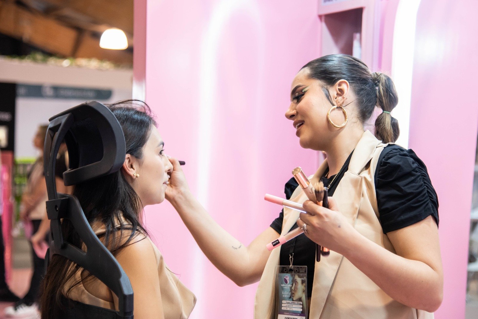 Maquilladora aplica maquillaje con una brocha a una mujer sentada en un stand de belleza con fondo rosa.