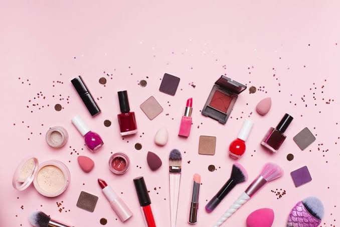 Productos de maquillaje, brochas, esponjas y esmaltes de uñas distribuidos sobre un fondo rosa con confeti brillante.